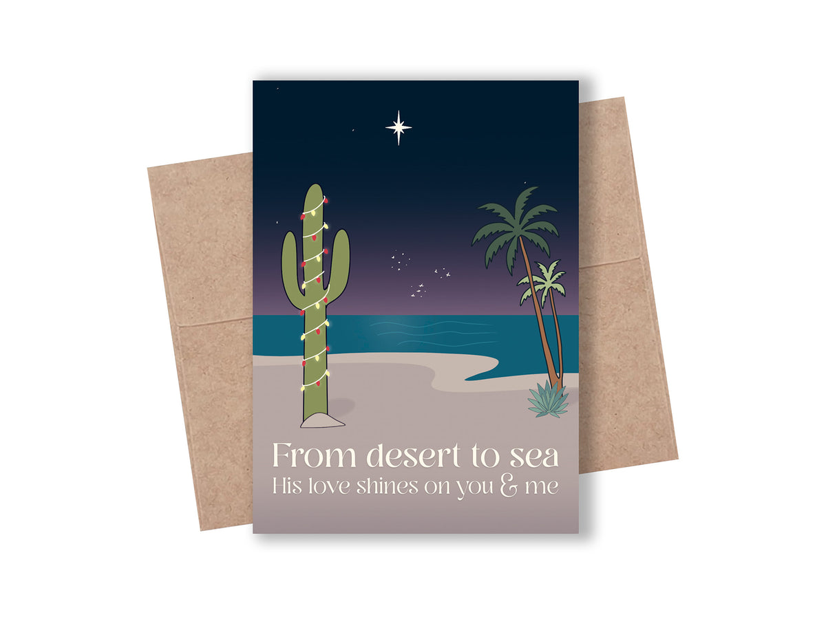 Christian beach Christmas card with a saguaro cactus wrapped in string lights, palm trees, and a bright star over the ocean at night. Text reads “From desert to sea, His love shines on you & me.” Shown with a kraft envelope.