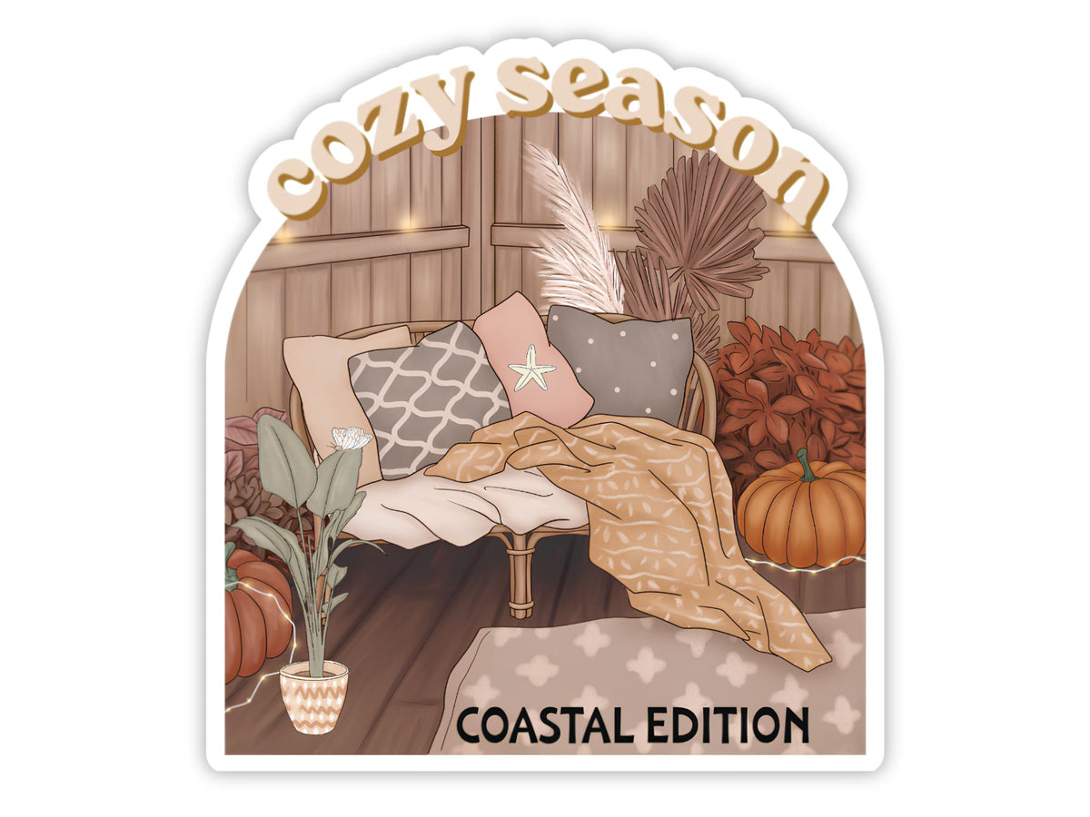 "Cozy Season" cute pumpkin sticker, Coastal Edition. A rattan daybed with a starfish pillow and throw blanket, pumpkins, pampas grass, and dried palms under string lights