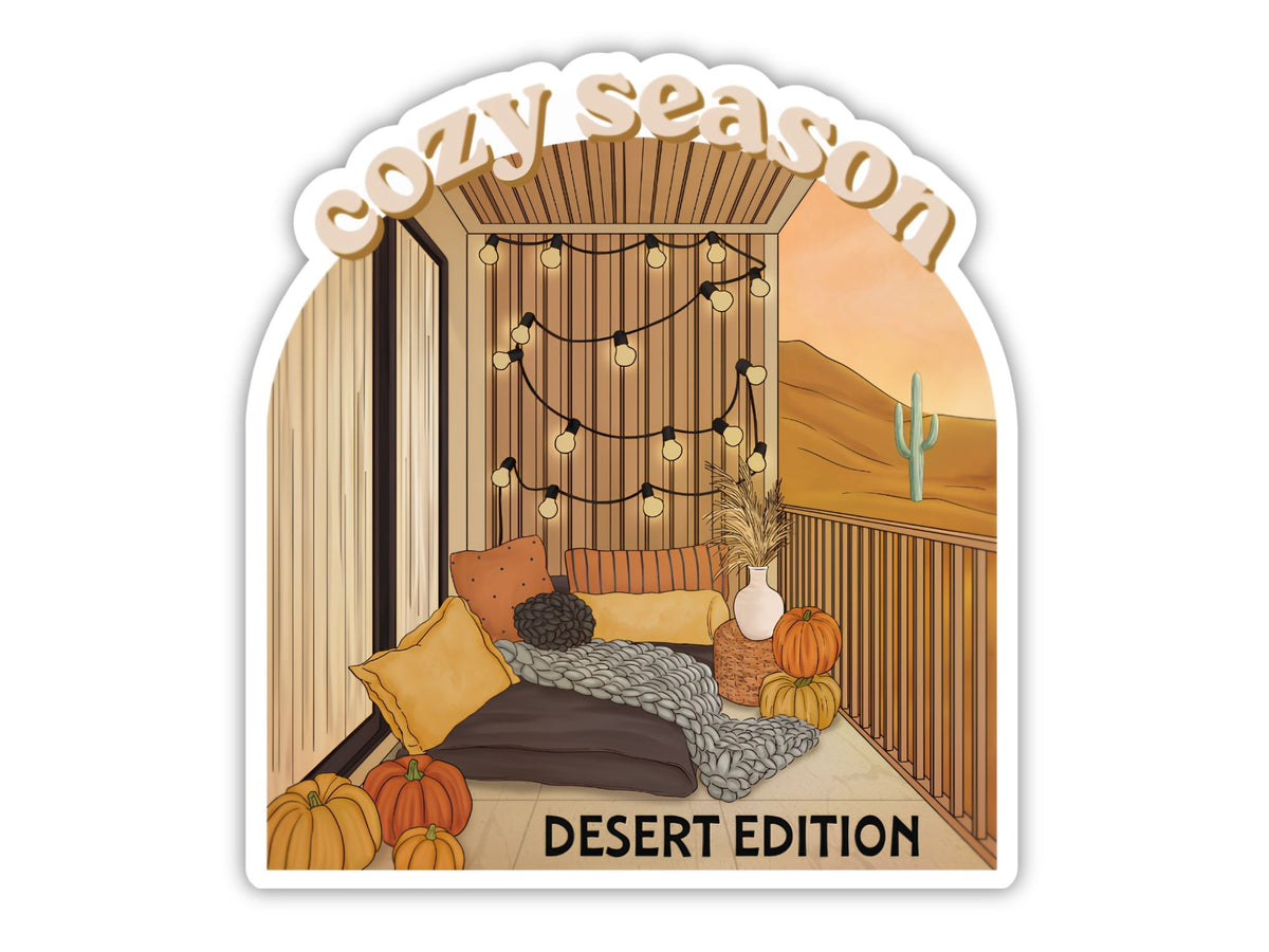 "Cozy Season" cute fall sticker, Desert Edition. A balcony strung with lights, pumpkins, a chunky knit blanket over floor cushions, and a lone saguaro against golden hills at sunset