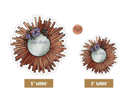 "Stop. Look. Smile." self love sticker in 5-inch and 3-inch sizes next to a penny for scale, copper sunburst mirror with purple flowers.