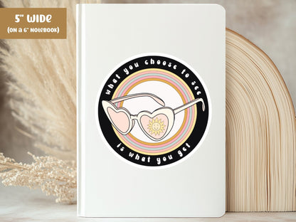 Notebook with a motivational sticker featuring sunglasses and a heart, with text 'What you choose to see is what you get', on a neutral background.